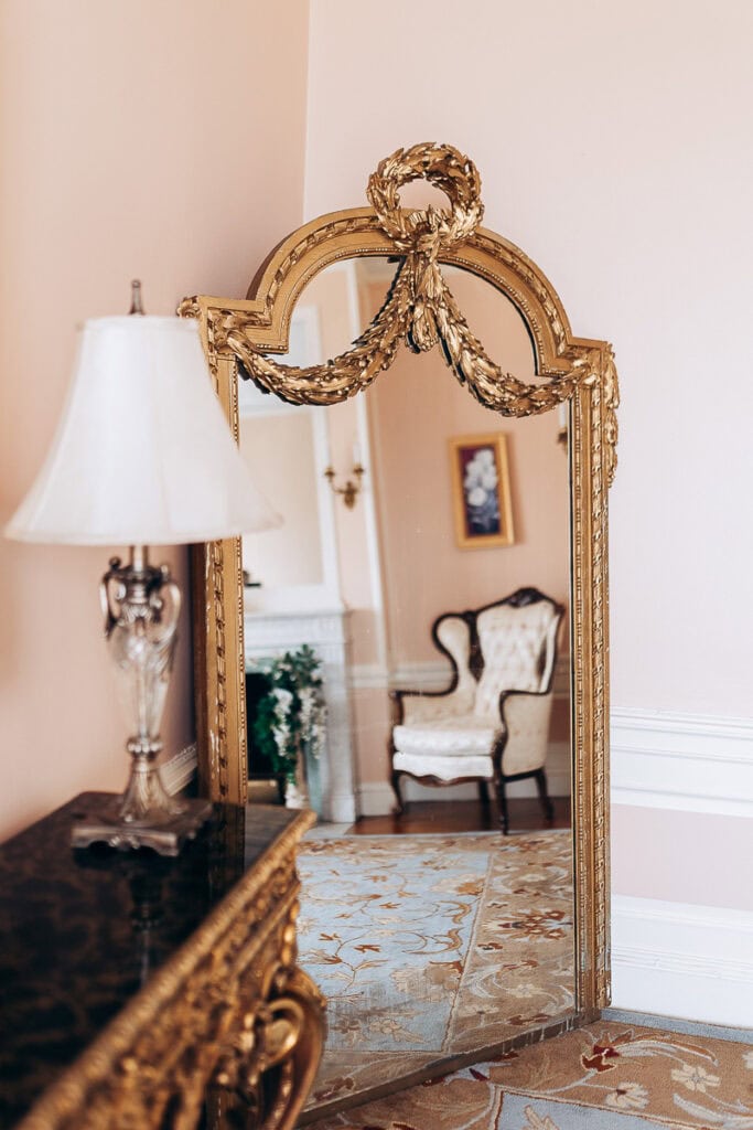 Gilded ornate mirror reflecting a tufted vintage armchair and floral rug in an elegant room with soft pink walls, New York, NY.