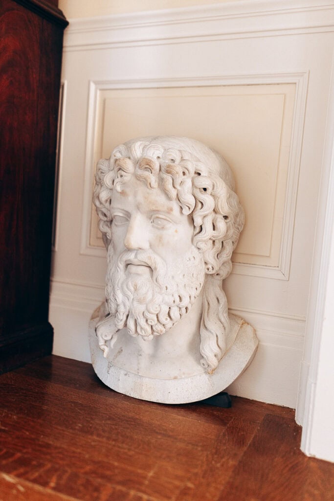 Marble bust of a bearded man with curly hair placed on wooden floor against cream paneled wall in an elegant interior.