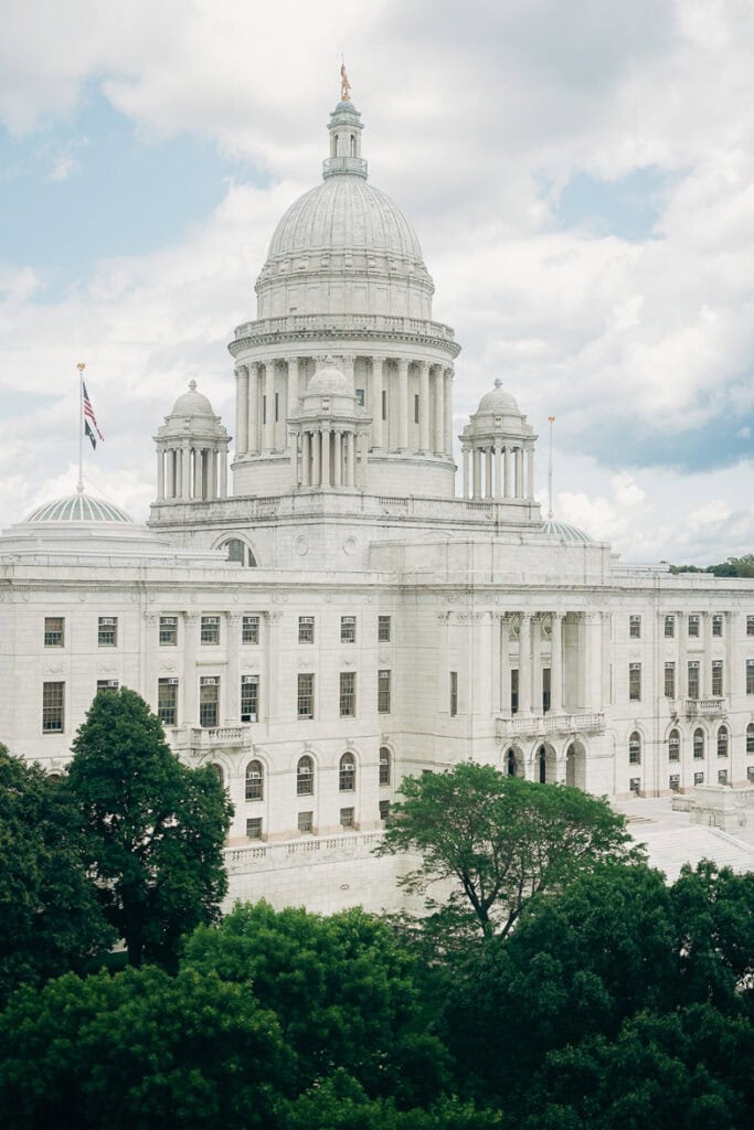 Neoclassical state capitol building with large dome, columns, and American flag, surrounded by dense green trees under a partly cloudy sky.