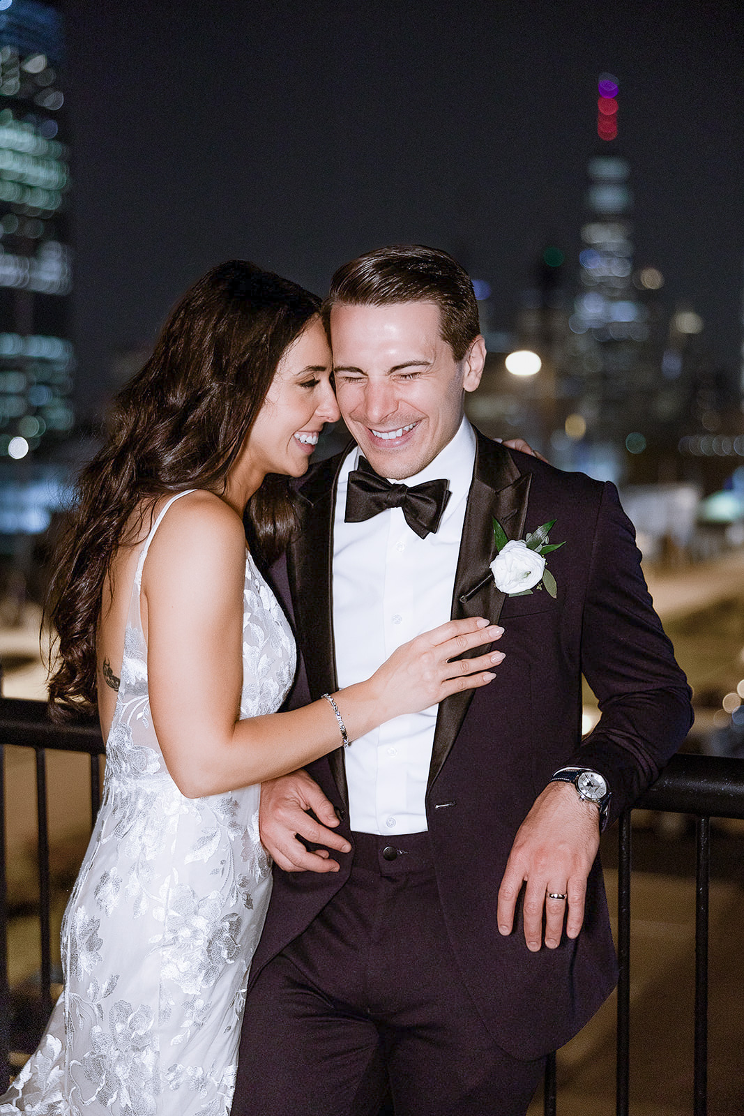 Bride in floral lace gown and groom in black tuxedo with bow tie, sharing a joyful moment at night with Manhattan skyline blurred in background.