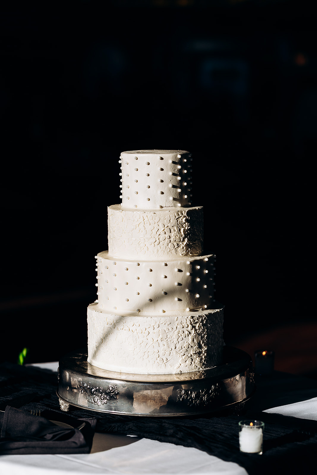 Four-tier white wedding cake with floral embossing and pearl dot details on a silver stand, lit dramatically against a dark background.