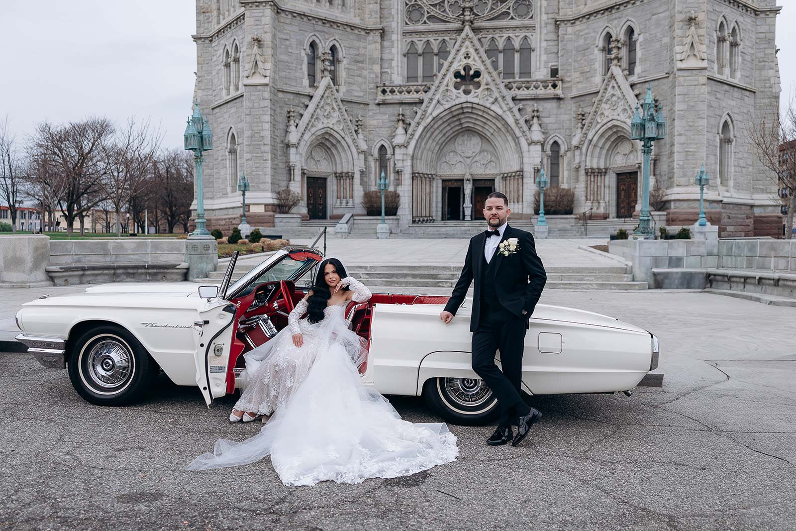 Bride in lace wedding dress sitting in a vintage white convertible with red interior, groom in black tuxedo leaning on car door, Gothic cathedral facade in background.