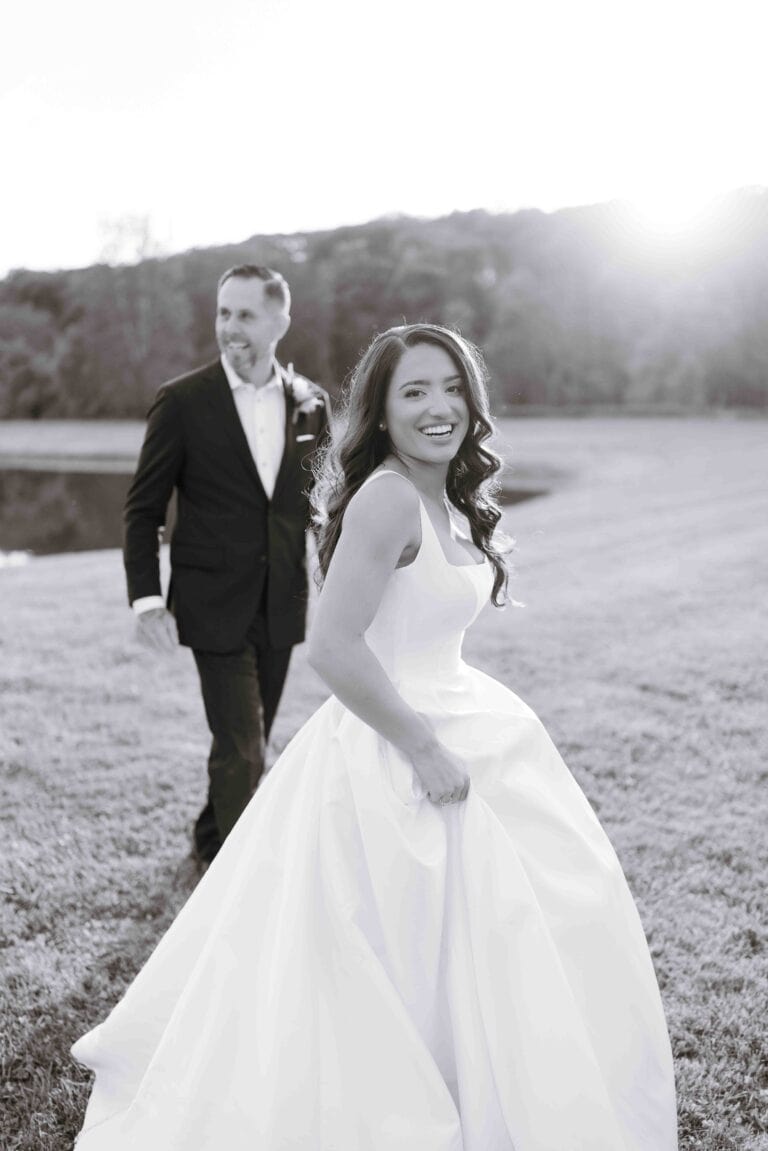 Black and white photo of a bride in a structured white gown smiling towards the camera with a groom in a black tuxedo blurred in the background outdoors.