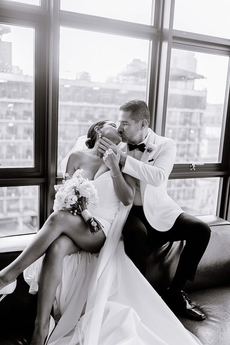 Bride and groom seated by large windows at Wythe Hotel, Brooklyn, sharing a kiss; bride holds white flower bouquet, groom in white tuxedo jacket.