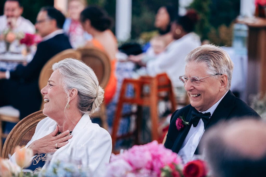 Elderly man in tuxedo and woman in white blazer laughing together at a wedding reception with floral centerpieces and guests in soft focus background.