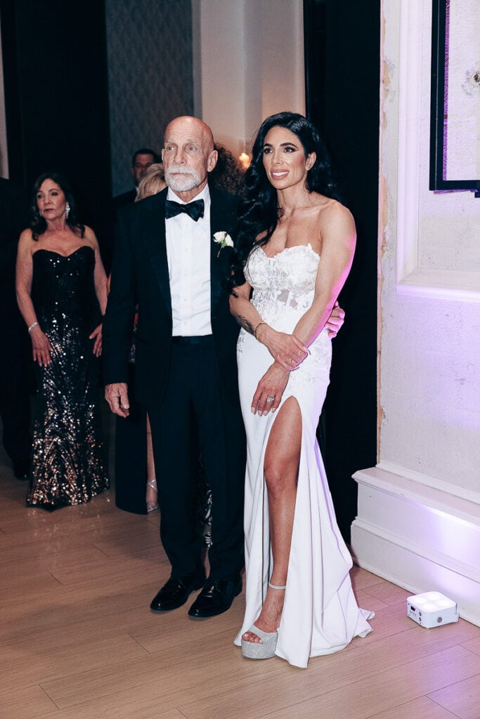 Bride in strapless white lace gown with thigh slit and platform heels standing next to an elder man in a black tuxedo at an indoor wedding venue in New York, NY.