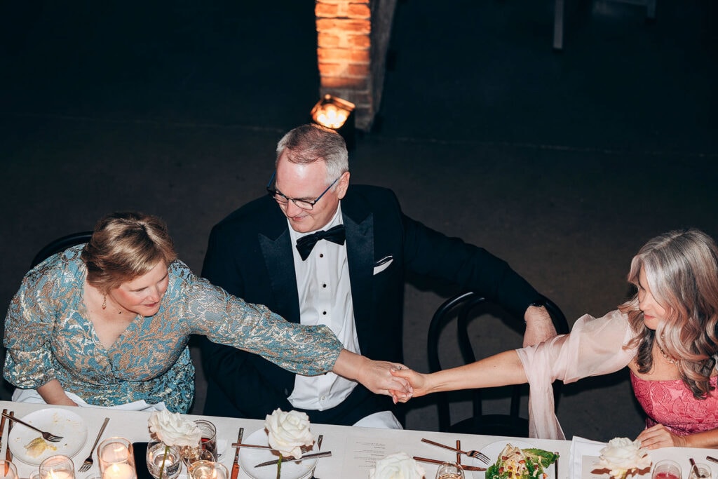 Three guests at a wedding reception in New York, NY, seated at a table with white roses and candles, two women holding hands across the table while a man in a tuxedo smiles between them.
