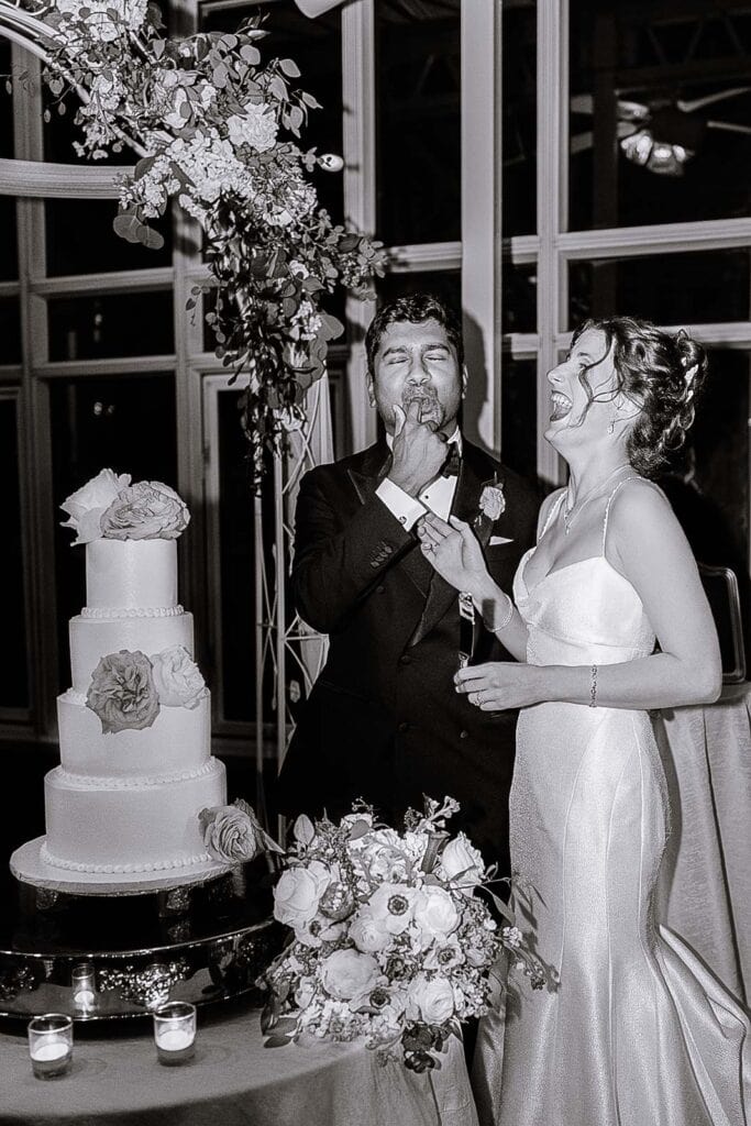 Groom tasting wedding cake with finger as bride laughs beside him near floral arrangement and three-tier cake in a Manhattan wedding venue.