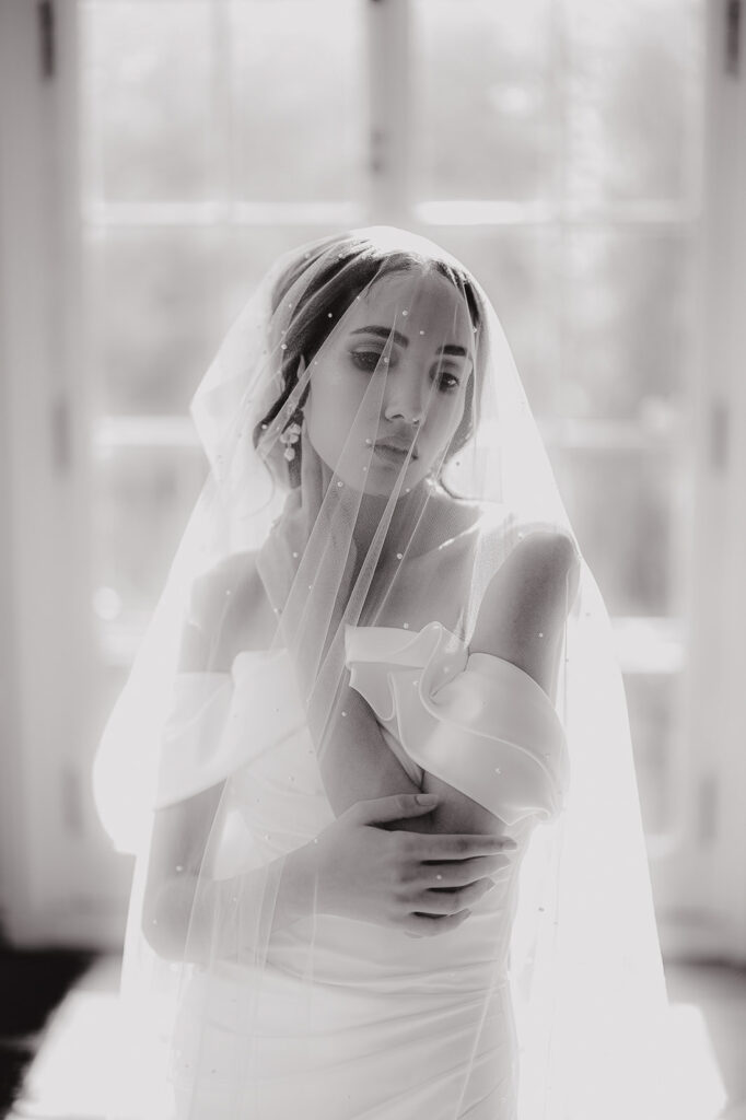 Black and white portrait of a bride in an off-shoulder wedding dress with a sheer, beaded veil softly illuminated by natural light through a window.