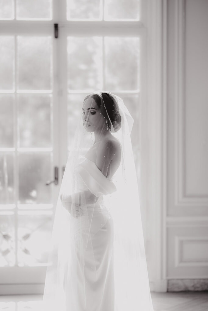 Side profile of a bride in an off-shoulder wedding dress and pearl-dotted veil standing near a large window with soft natural light in New York, NY.