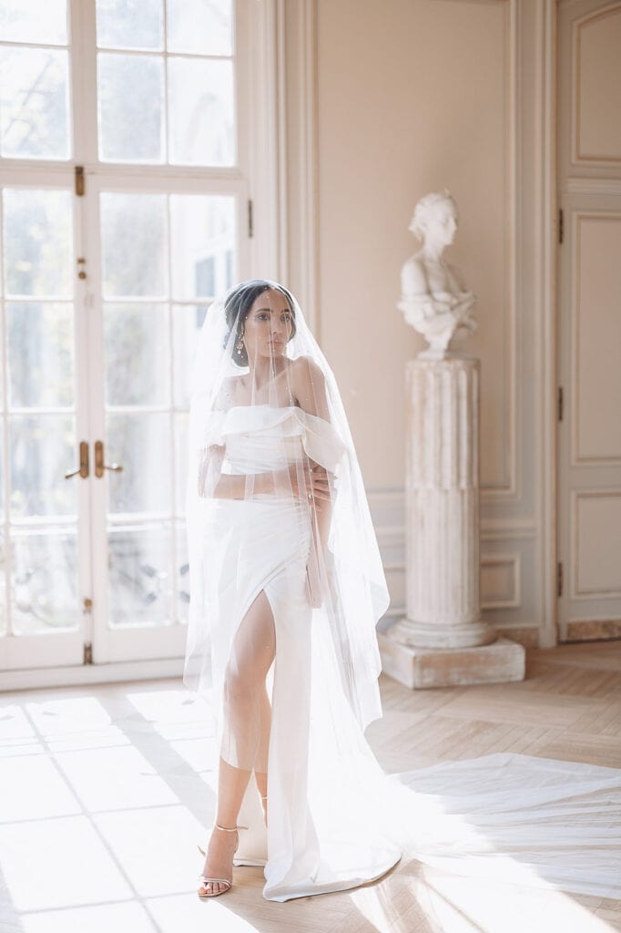 Bride in off-shoulder white gown with thigh slit and sheer veil standing in bright room with large windows and classical sculpture in New York, NY.