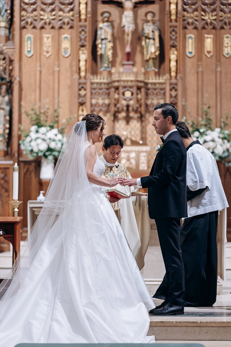 Bride in flowing white gown and veil placing ring on groom's finger during wedding ceremony at ornately decorated church altar with priest and assistant.