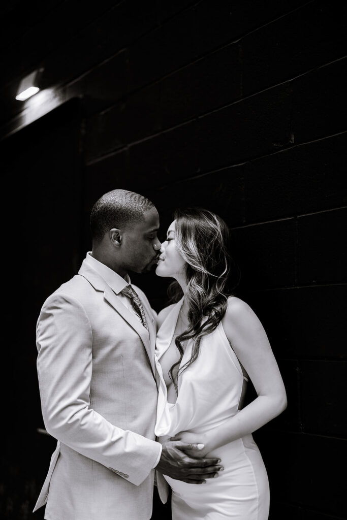 Black and white photo of a couple kissing, the man in a light suit holding the woman's pregnant belly, standing against a dark brick wall in New York.