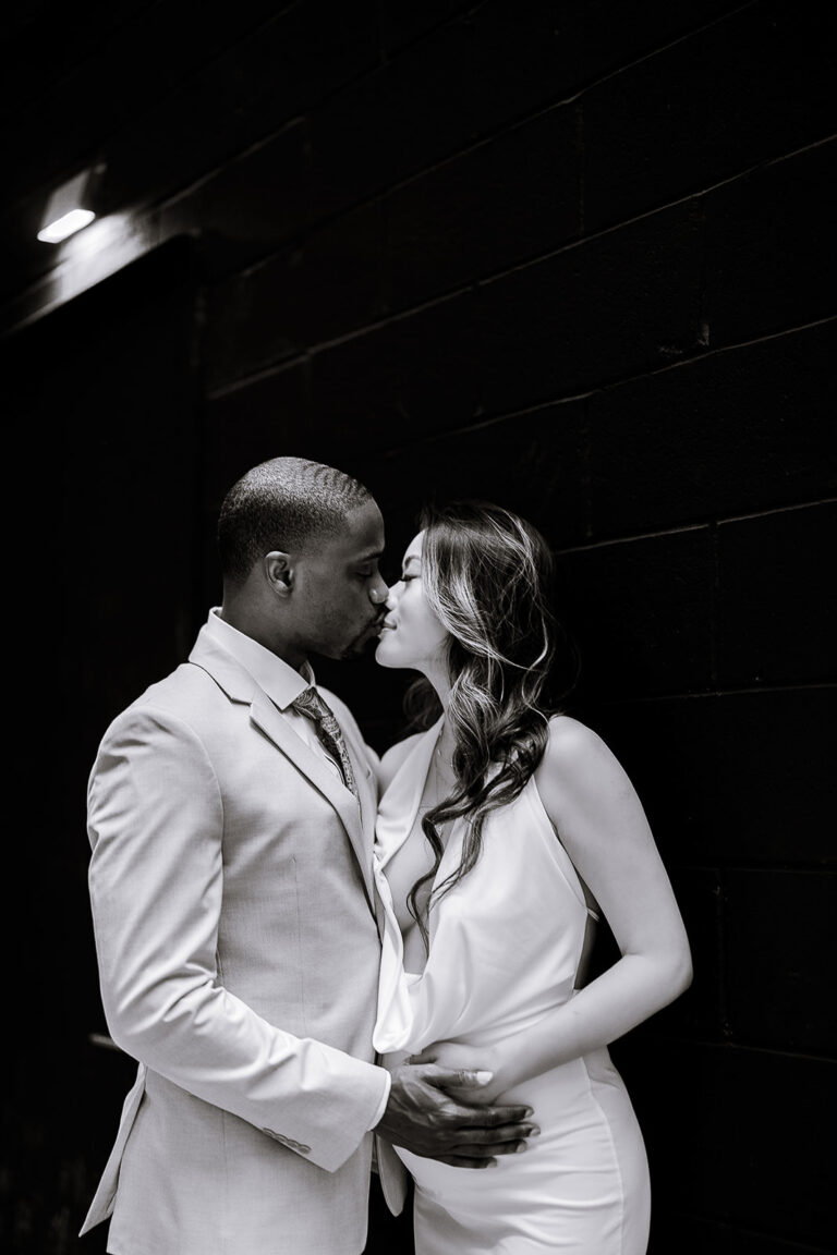 Black and white photo of a couple kissing, the man in a light suit holding the woman's pregnant belly, standing against a dark brick wall in New York.
