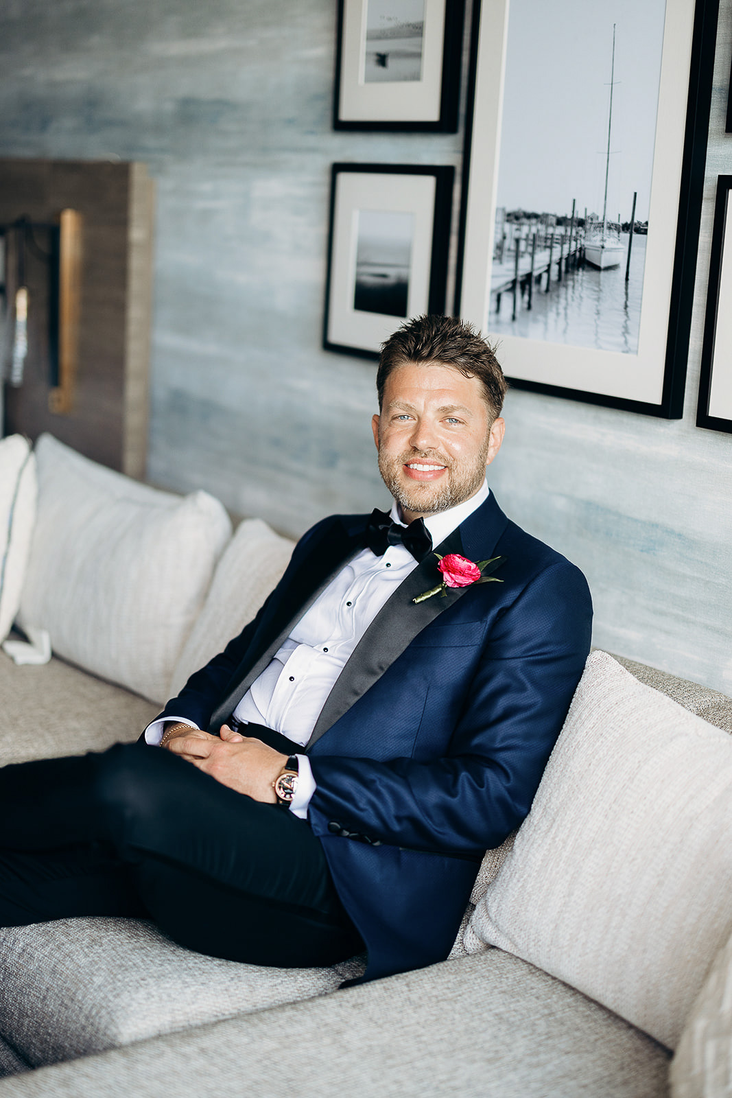 Man in navy tuxedo with black satin lapel and red rose boutonniere sitting on beige sofa against wall with black and white framed photos in New York, NY.