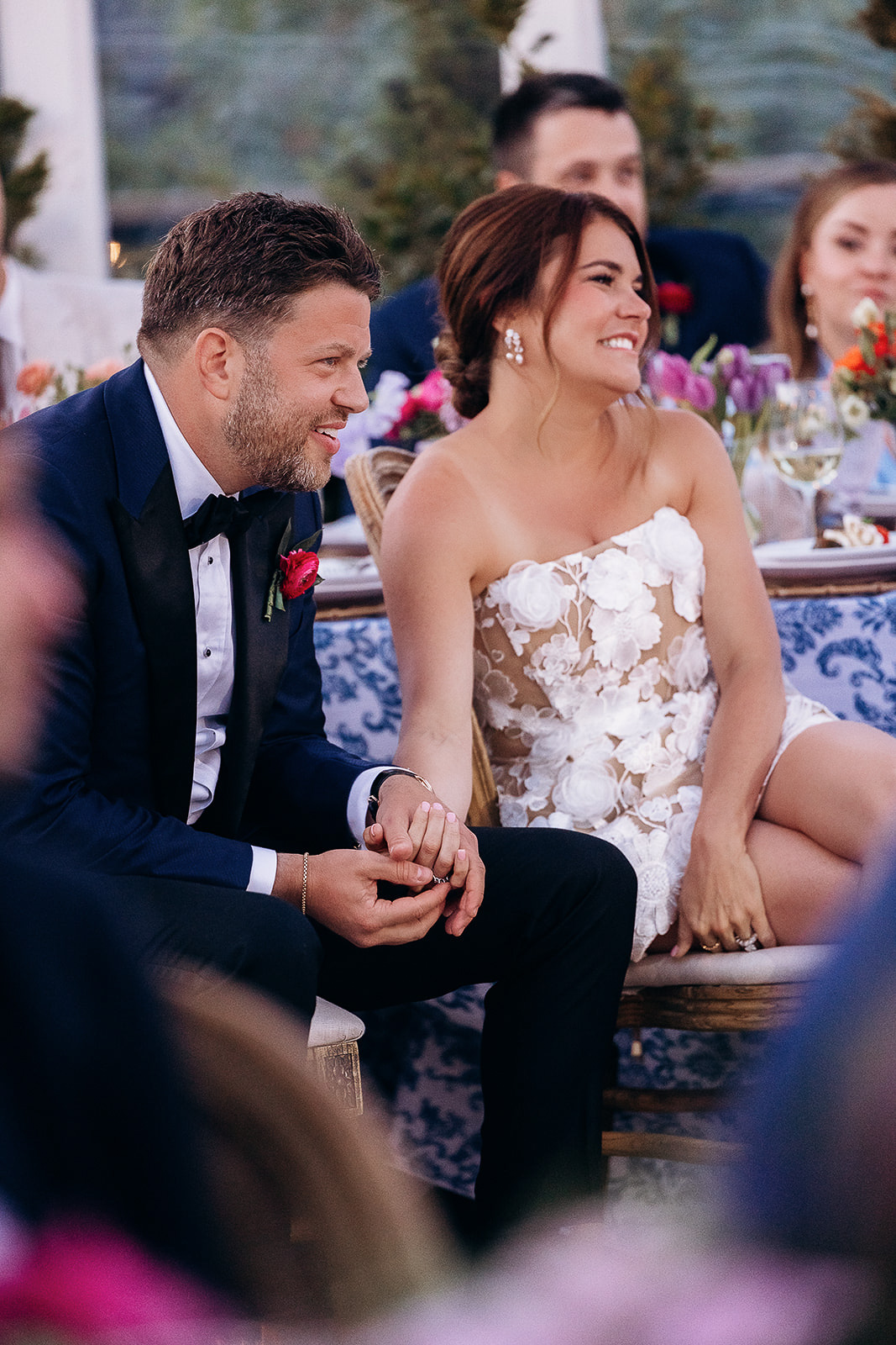 Newlywed couple seated closely, holding hands during wedding reception with floral decor and guests in soft focus in New York, NY.
