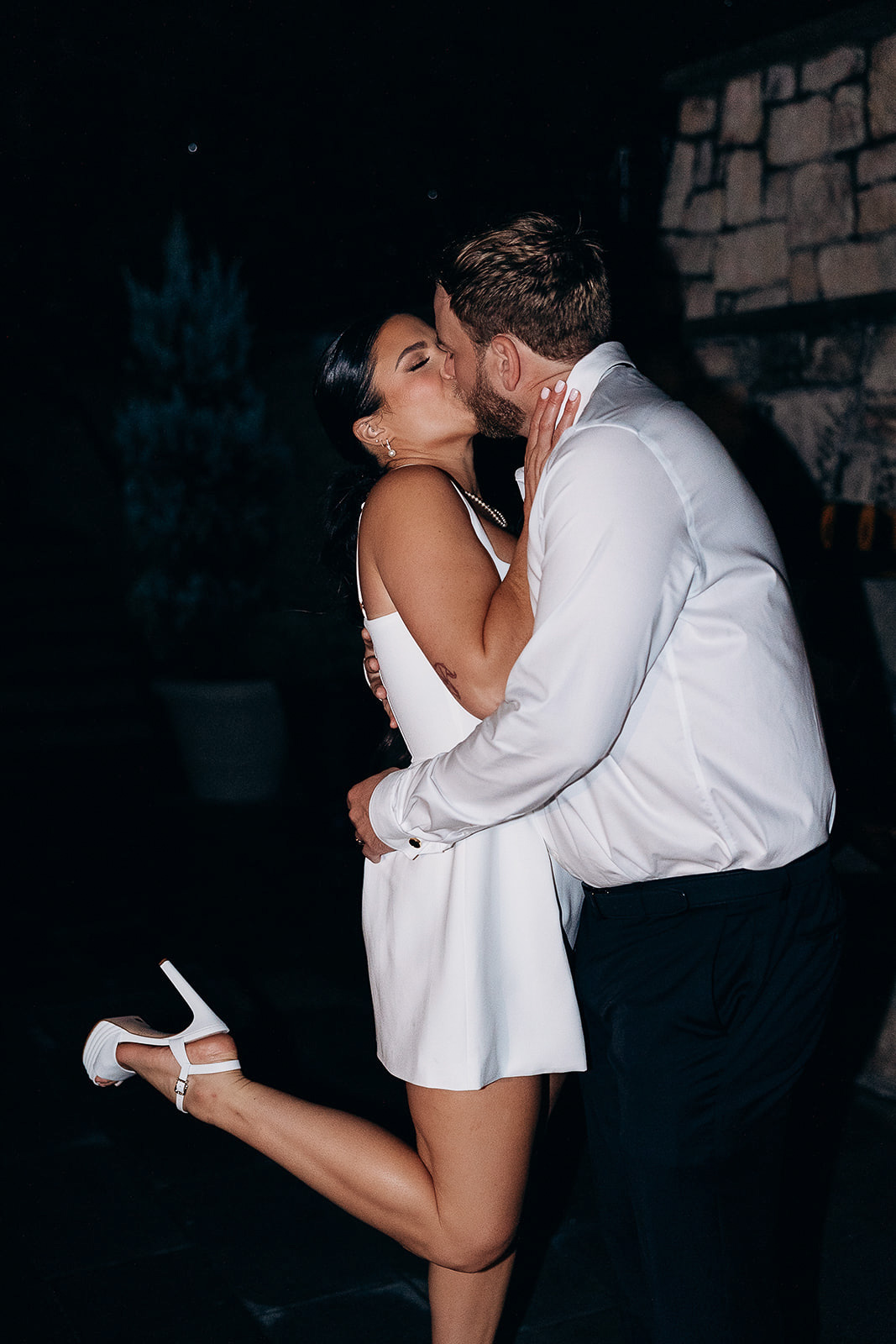 Couple embracing and kissing at night near a stone wall, woman in white dress with pearl necklace, man in white shirt, New York, NY.