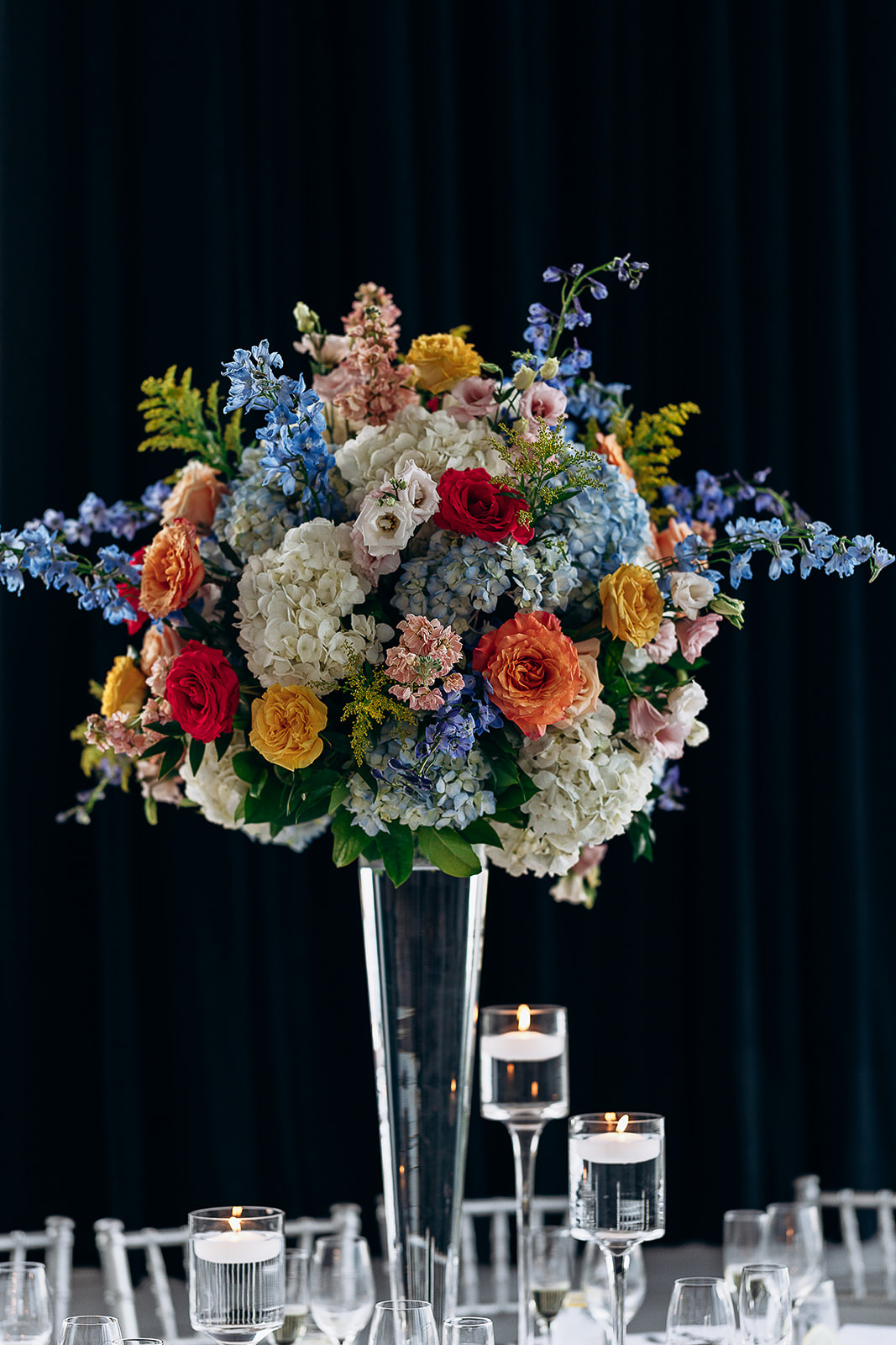 Tall crystal vase holding a sculptural floral arrangement with white hydrangeas, orange and red roses, blue delphiniums, and greenery, surrounded by floating candles and glassware on a white table.