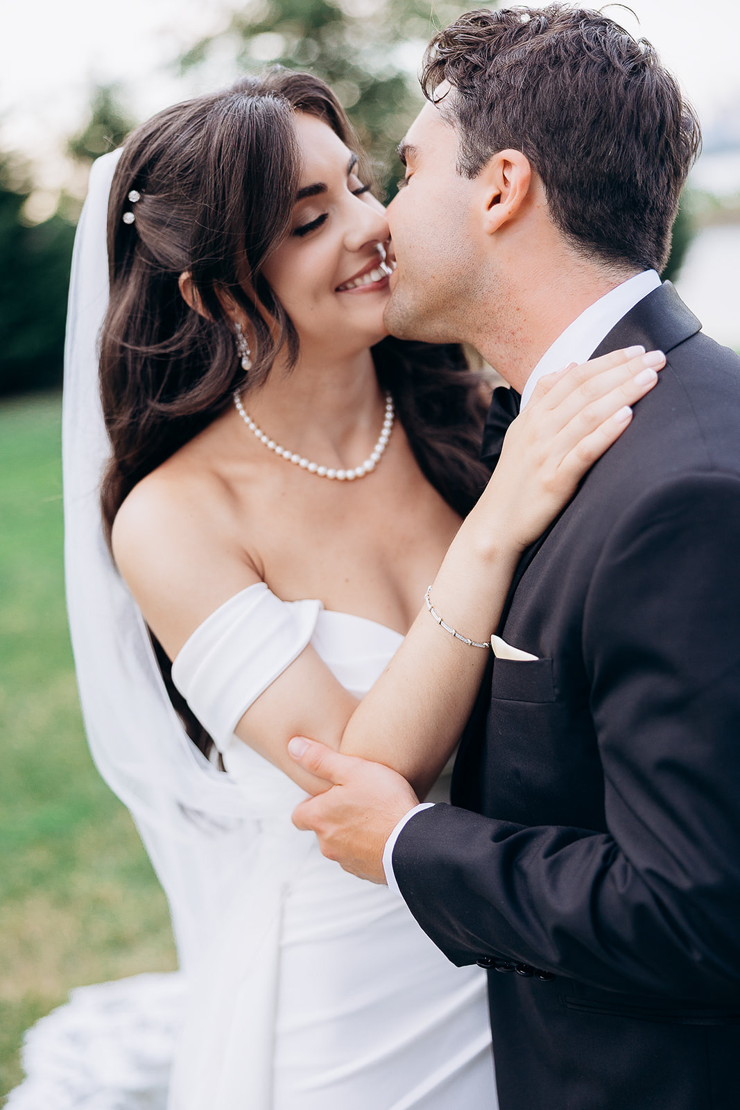 Bride in off-shoulder white wedding dress and pearl necklace gently holding groom in black tuxedo as they lean in for a kiss outdoors in New York, NY.