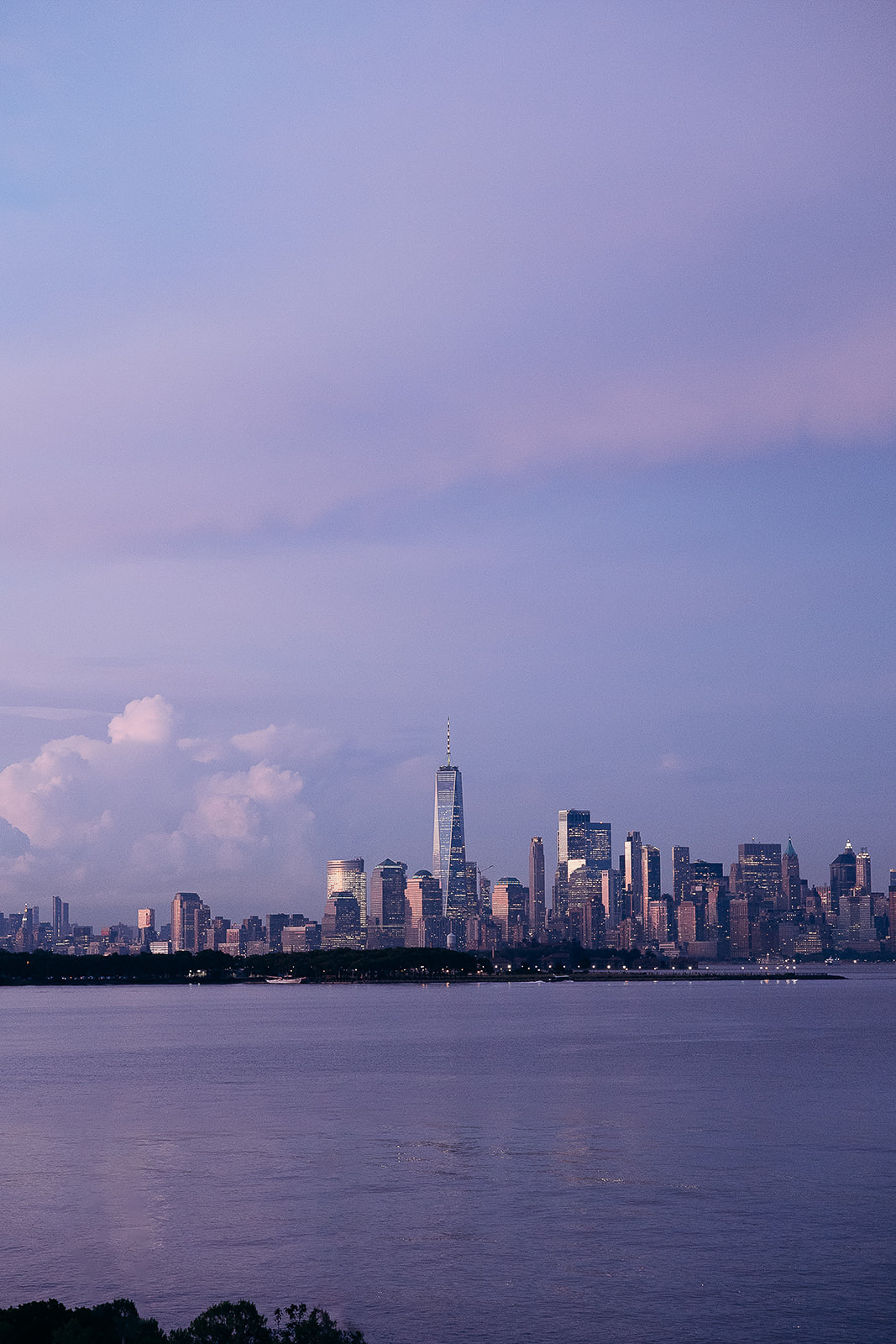 View of Manhattan skyline at twilight featuring One World Trade Center and surrounding skyscrapers with calm water in foreground in New York, NY.