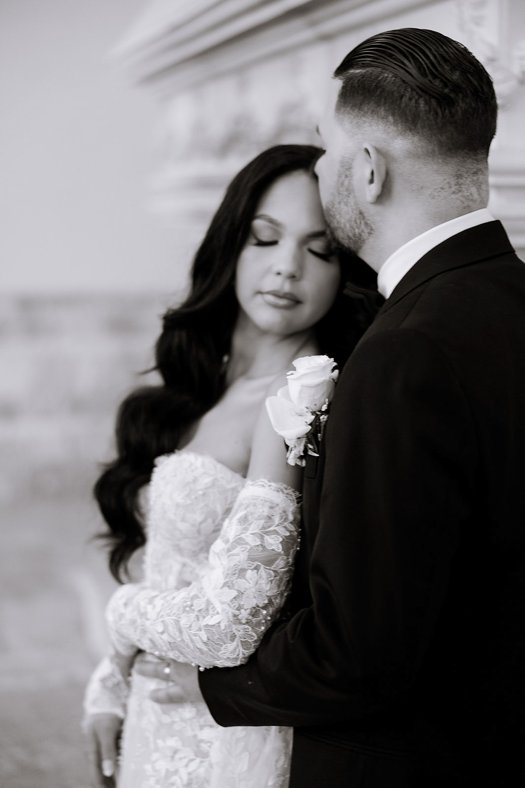 Bride in lace wedding gown with long sleeves embraces groom in black suit with white boutonniere, eyes closed in a tender moment in New York, NY.