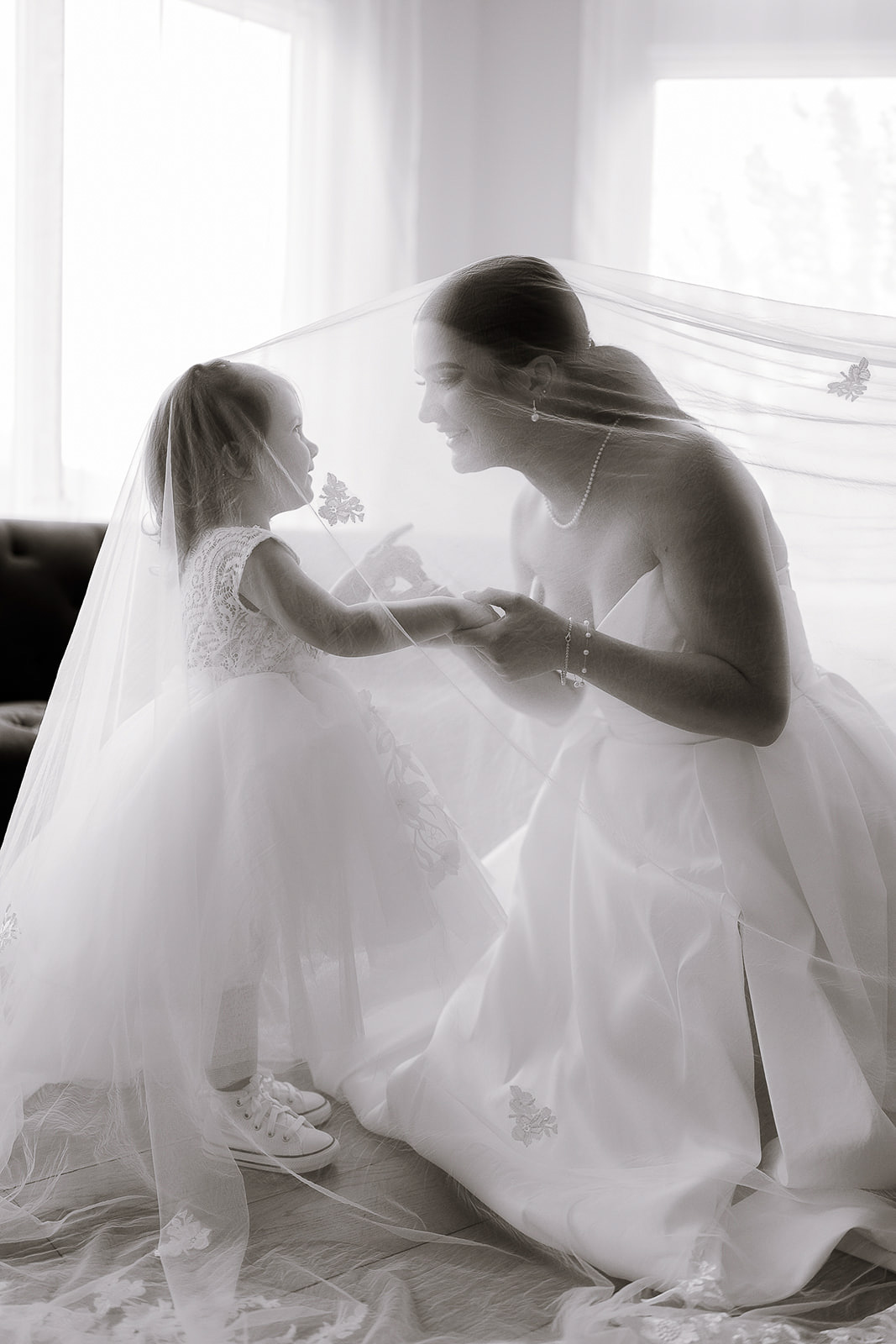 Bride and young flower girl sharing a moment under a sheer bridal veil in a softly illuminated room in New York, NY.