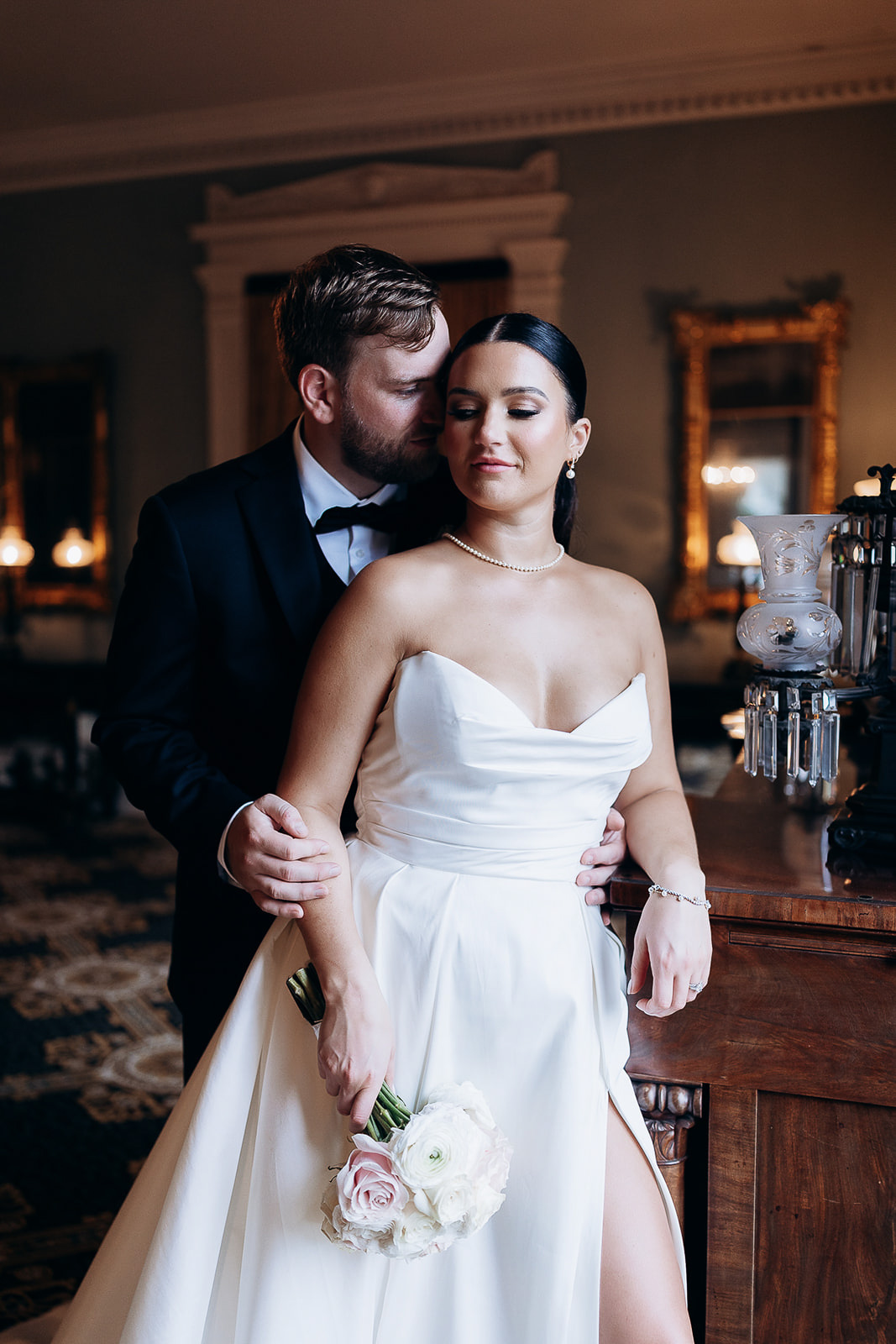 Bride in strapless satin wedding dress holding a bouquet of white and pale pink roses, embraced by groom in black tuxedo in an elegant room with antique furnishings and warm lighting.