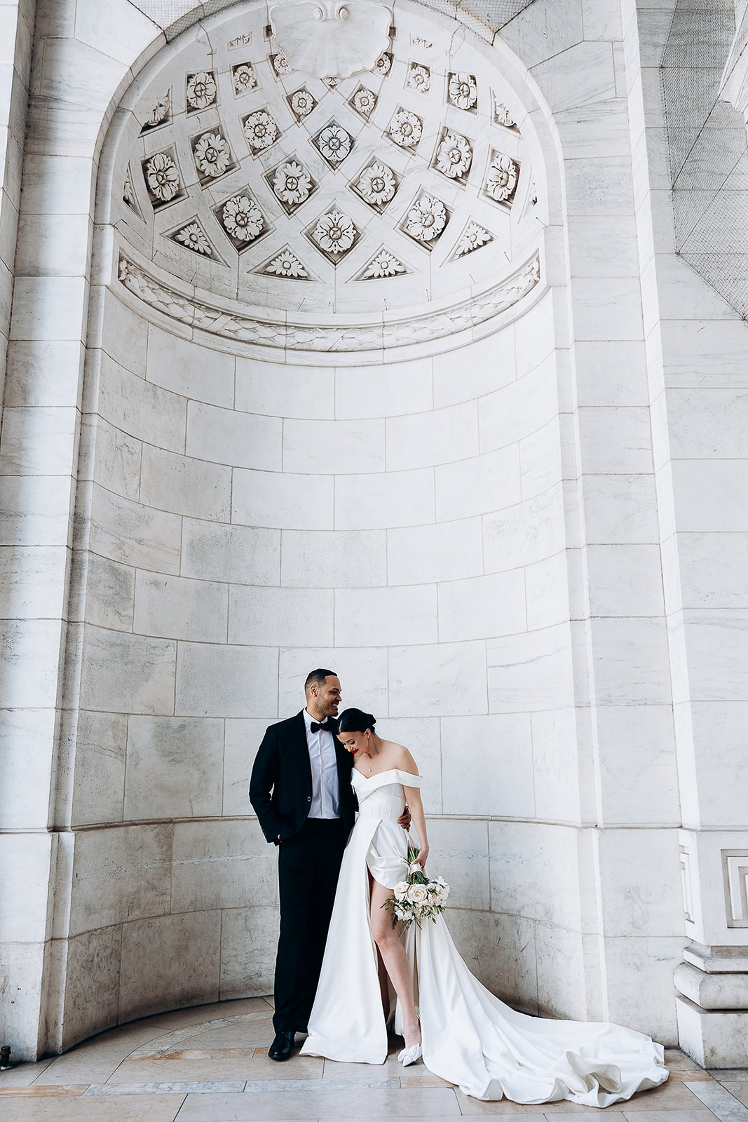 Bride in off-shoulder white gown holding bouquet, leaning into groom in black tuxedo, standing under ornate marble arch in New York, NY.