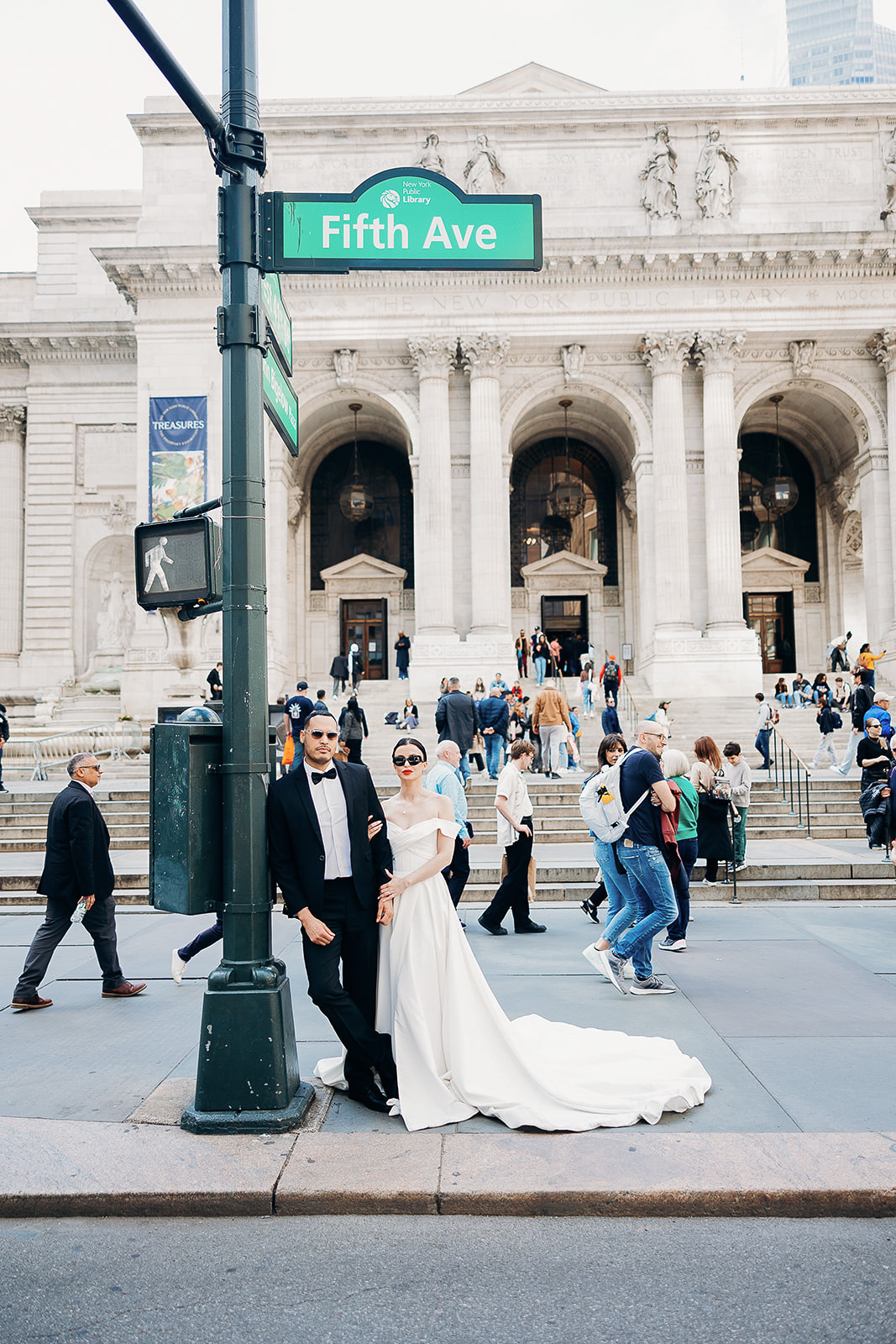 Bride in off-shoulder white gown and groom in black tuxedo standing by Fifth Avenue street sign with New York Public Library steps and crowd in background.