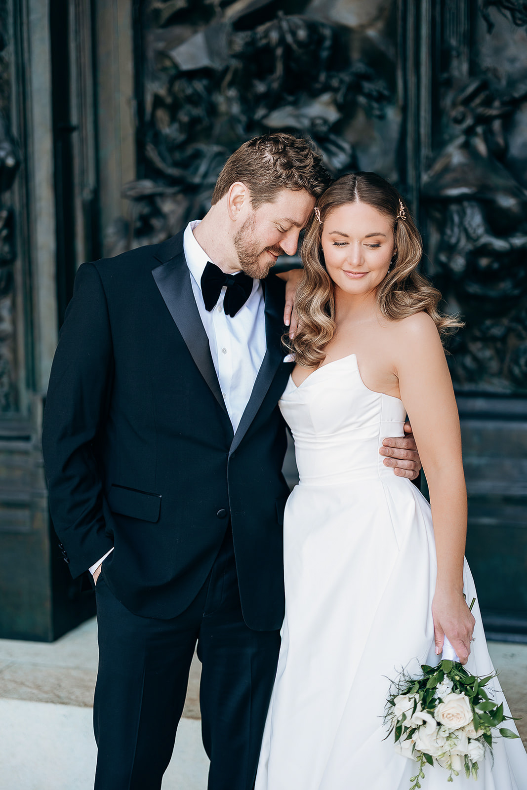 Bride in strapless white gown holding bouquet, groom in black tuxedo with bow tie, standing close with eyes closed near ornate dark sculptural wall in Manhattan, New York.