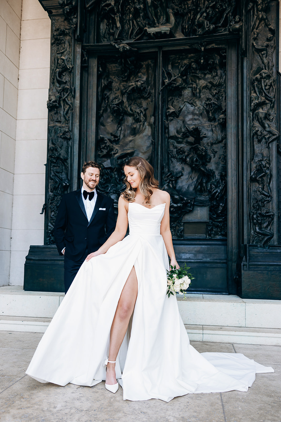 Bride in strapless white gown with thigh slit holding bouquet, groom in black tuxedo smiling, standing before ornate bronze sculpted doors in New York, NY.