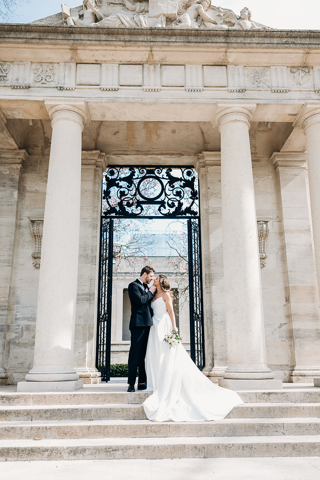 Bride in flowing white gown and groom in black tuxedo embrace on stone steps beneath neoclassical columns and ornate black iron gate in New York, NY.