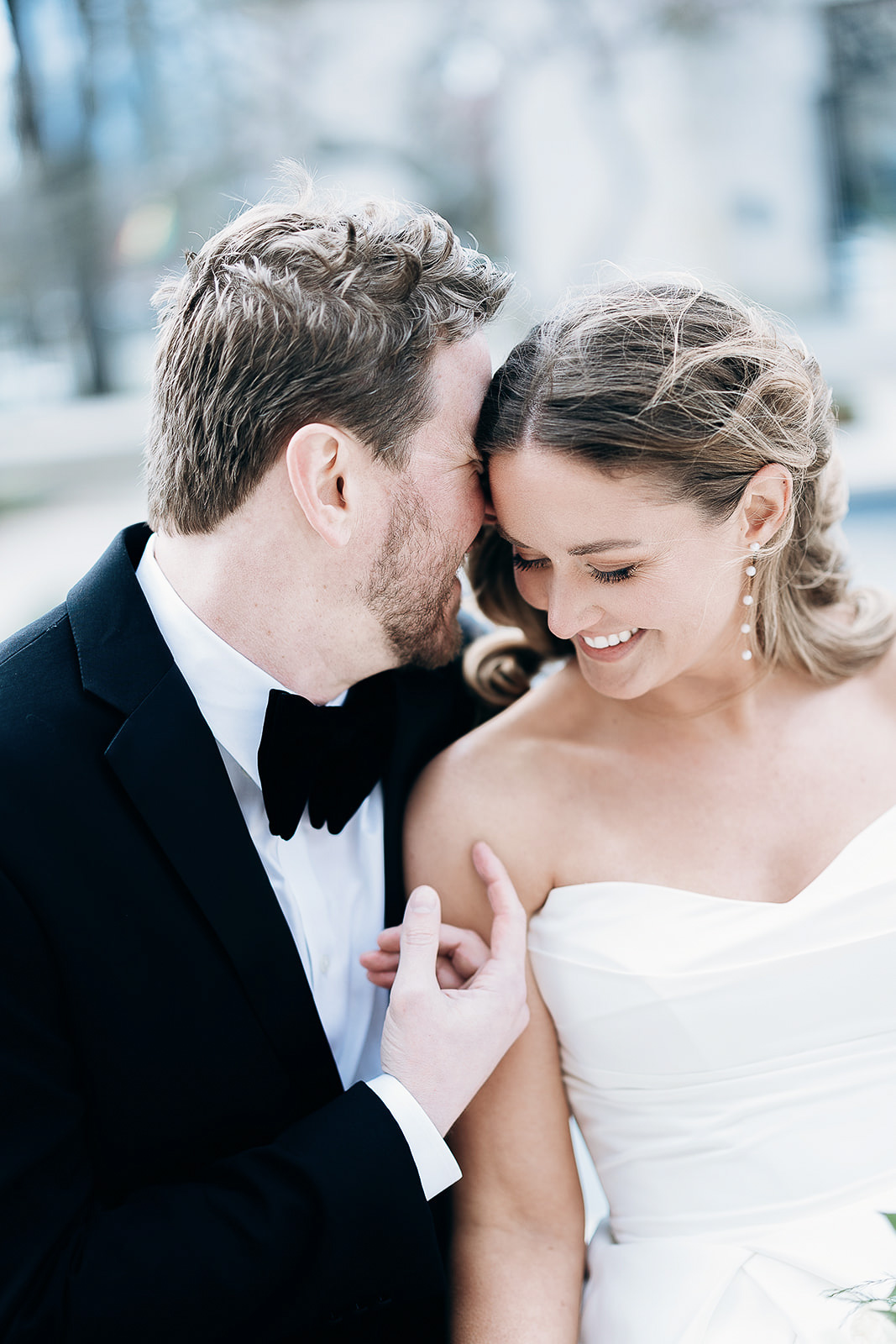 Close-up of groom whispering to bride, both smiling, groom in black tuxedo, bride in white off-shoulder dress with pearl earrings, outdoor setting.