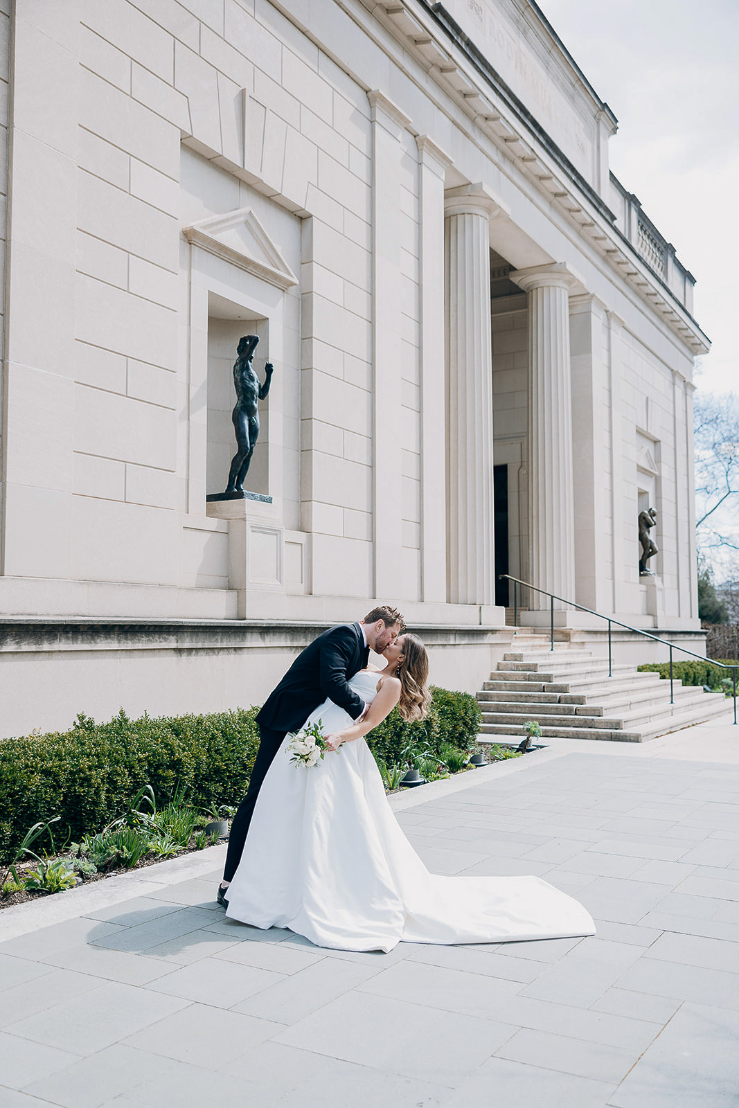 Bride in white gown and groom in black suit share a kiss while the groom dips the bride outside a neoclassical museum with large columns and statues in New York, NY.