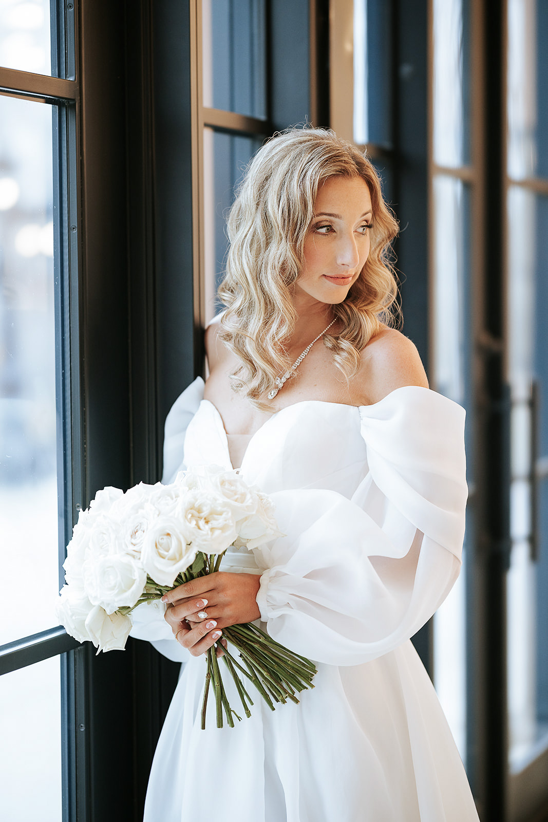 Bride in white off-shoulder wedding dress holding a bouquet of white roses, standing by large windows in New York, NY.