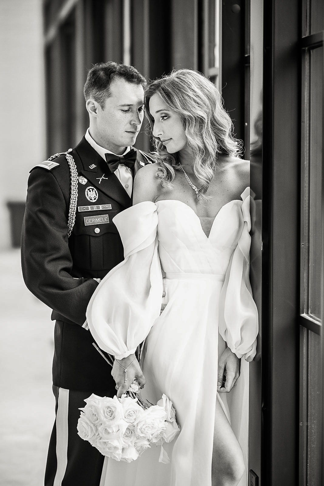 Black and white photo of a groom in military uniform embracing a bride in an off-shoulder wedding dress holding a bouquet of white roses by a window.