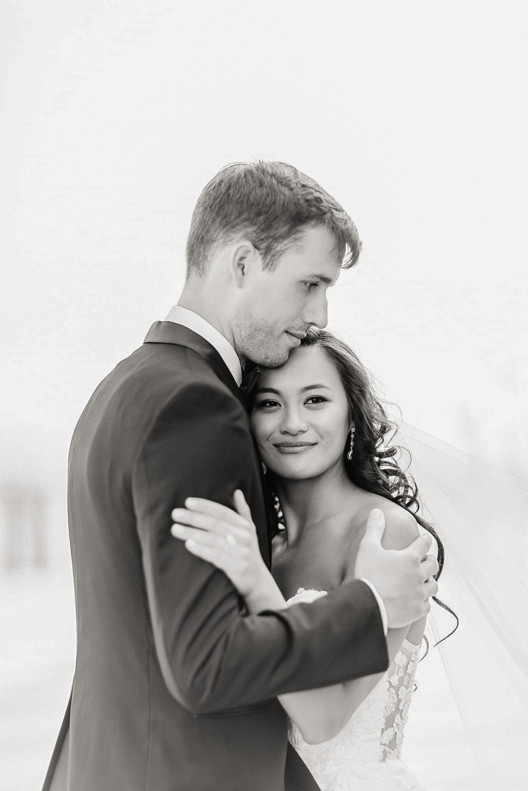 Black and white photo of a bride and groom embracing closely, the groom resting his head on the bride's forehead, both showing serene expressions.