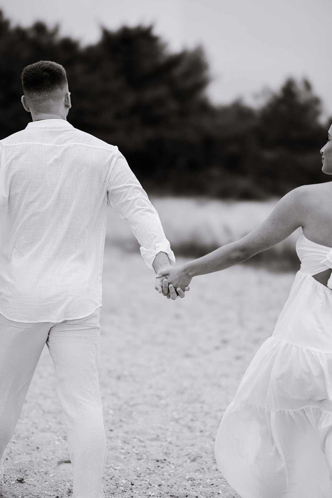 Couple holding hands walking away on a sandy path with blurred trees in the background, soft natural light, monochrome style.