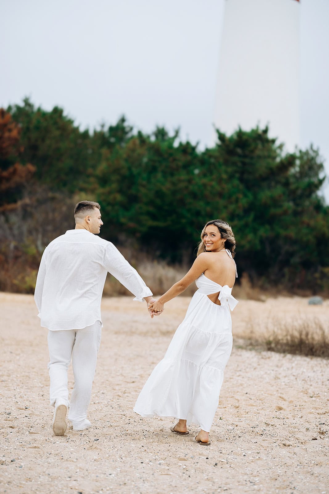 Couple walking hand-in-hand on sandy beach near Barnegat Light lighthouse, surrounded by green trees, woman in white dress looking back.