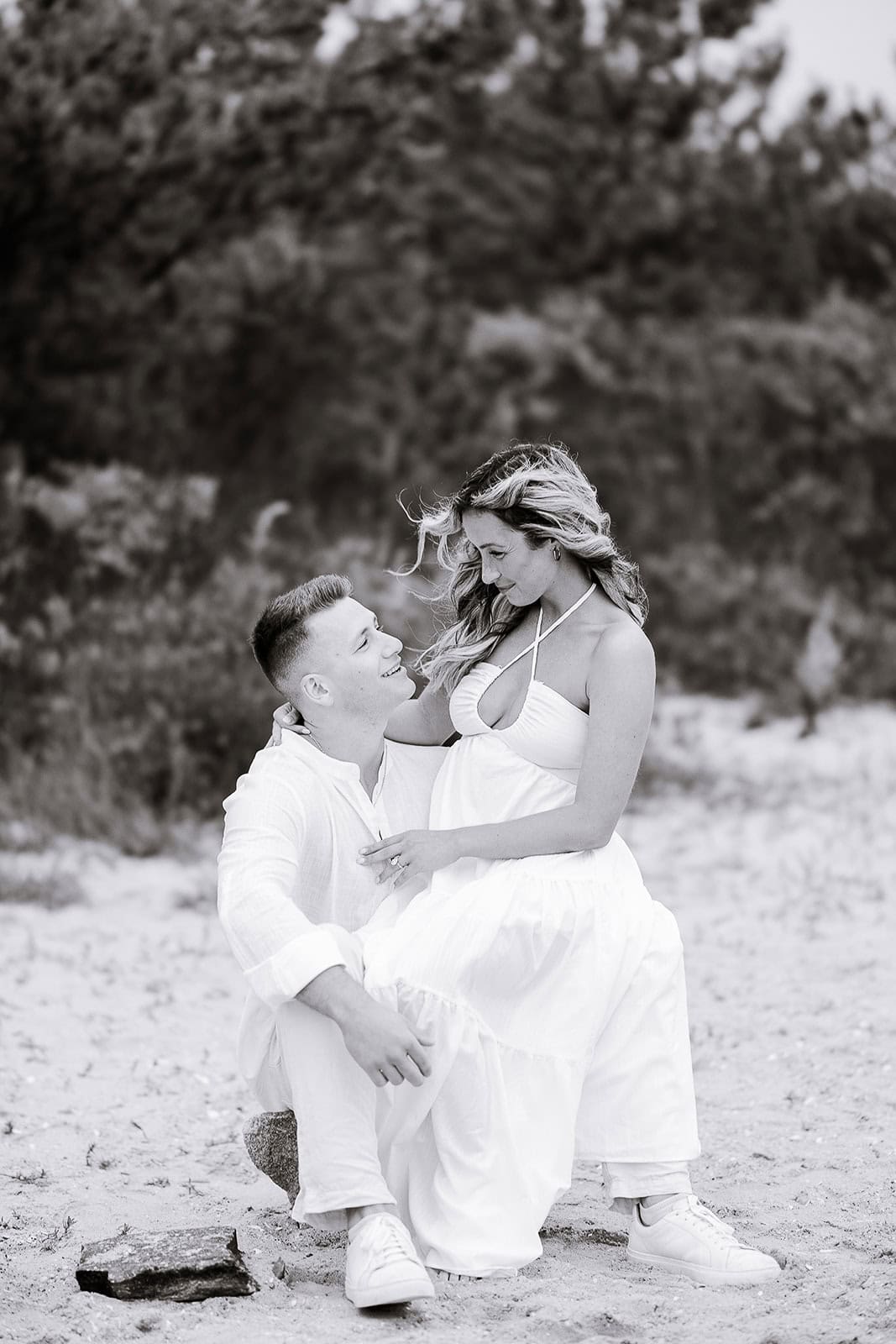 Black and white image of a couple on a sandy beach with trees in the background; the man sits while the woman sits on his knee, both dressed in white.