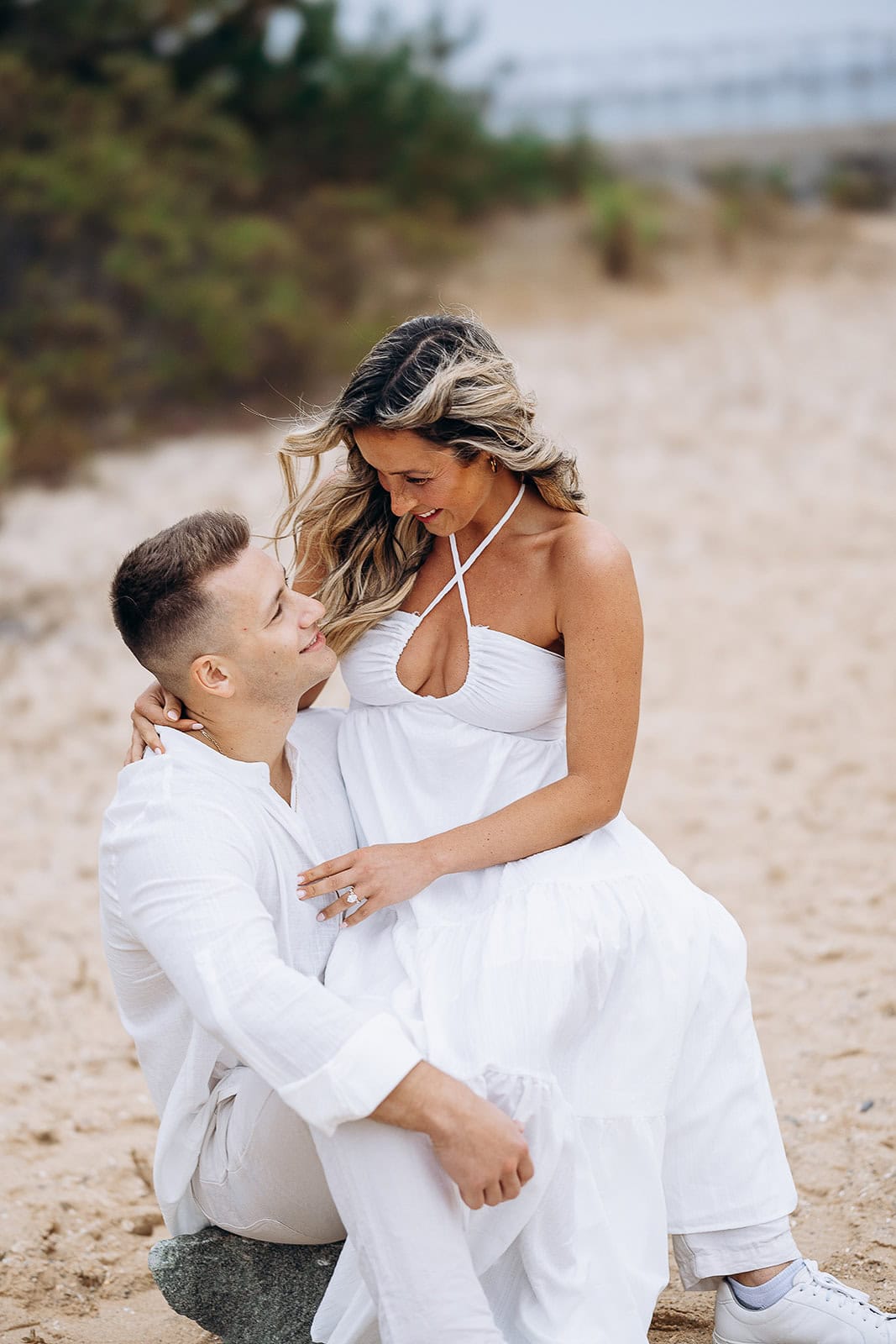 Couple dressed in white sitting on sand, woman seated on man's lap, both looking at each other with affection, sandy beach background.