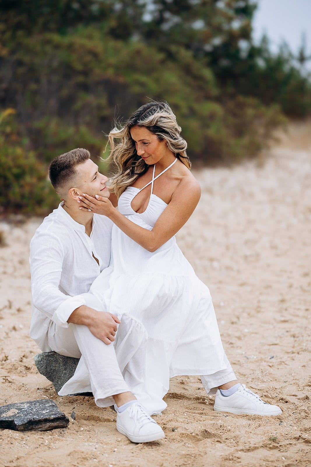 Couple dressed in white sitting on sandy beach with greenery in background, woman gently touching man's face in NJ.