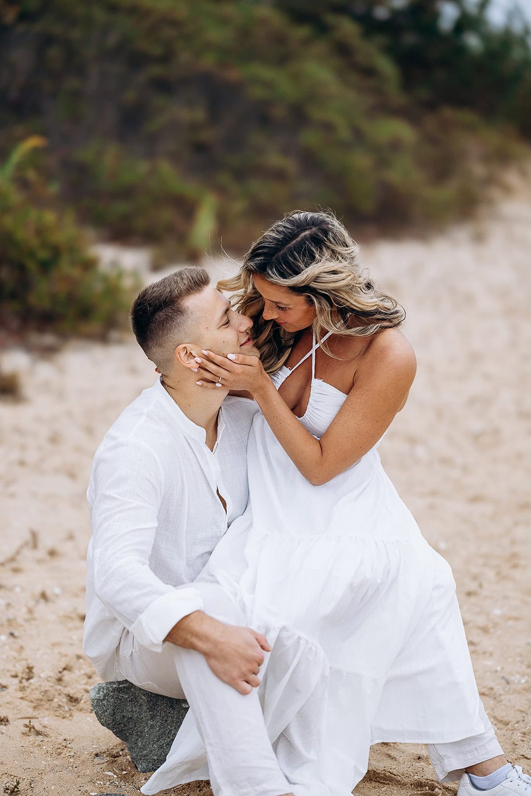 Couple dressed in white seated on sand, woman tenderly holding man's face, surrounded by blurred greenery in the background.