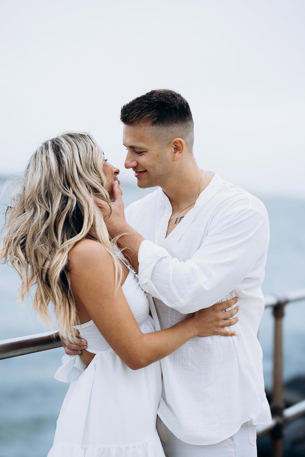 Couple in white embracing by the ocean, the man gently touching the woman's face, soft natural light, ocean and railing in background.