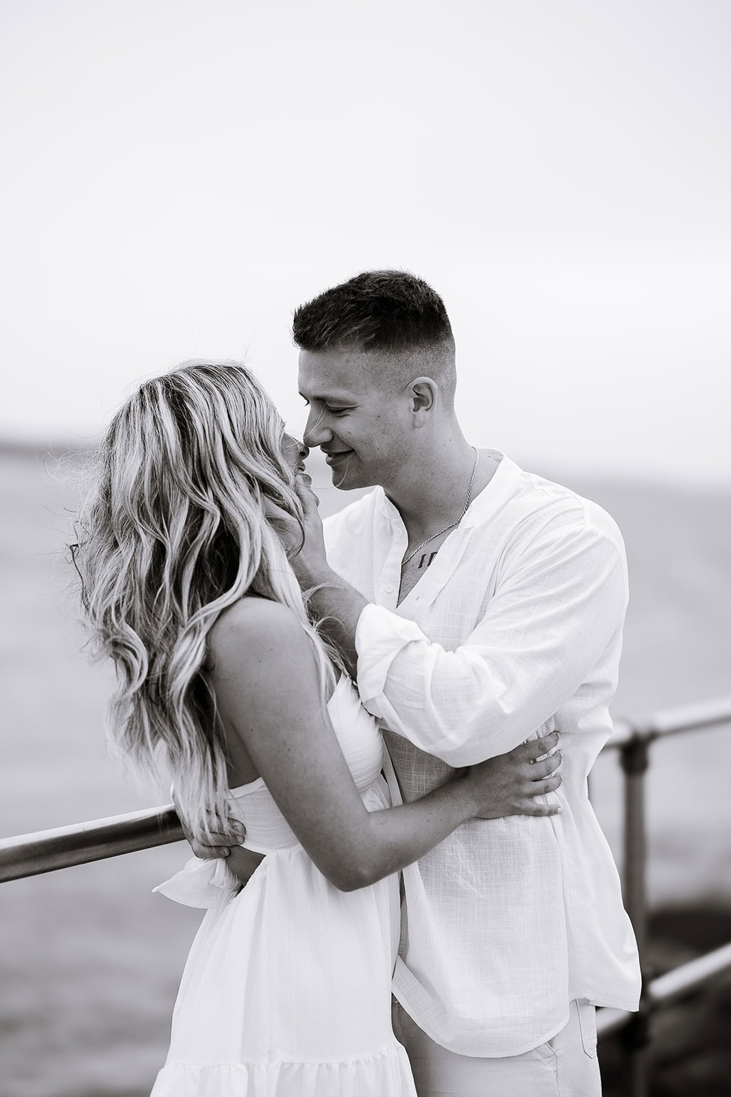Black and white image of a couple in white attire embracing closely near a waterfront railing, sharing an intimate moment.