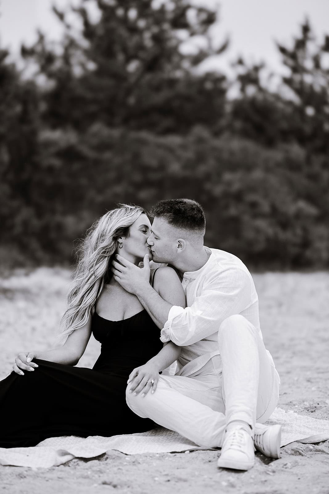 Black and white image of a couple sitting on a blanket outdoors, sharing a kiss with trees softly blurred in the background.