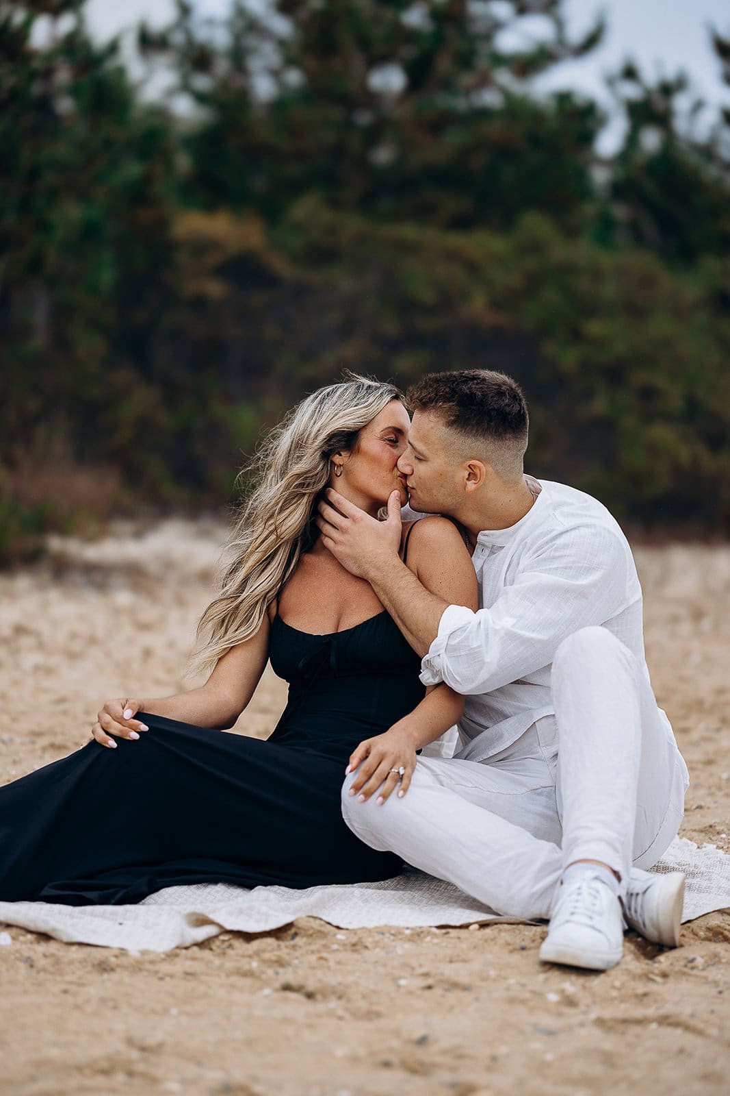 Couple sitting on a blanket on the beach, sharing a tender kiss with trees blurred in the background.