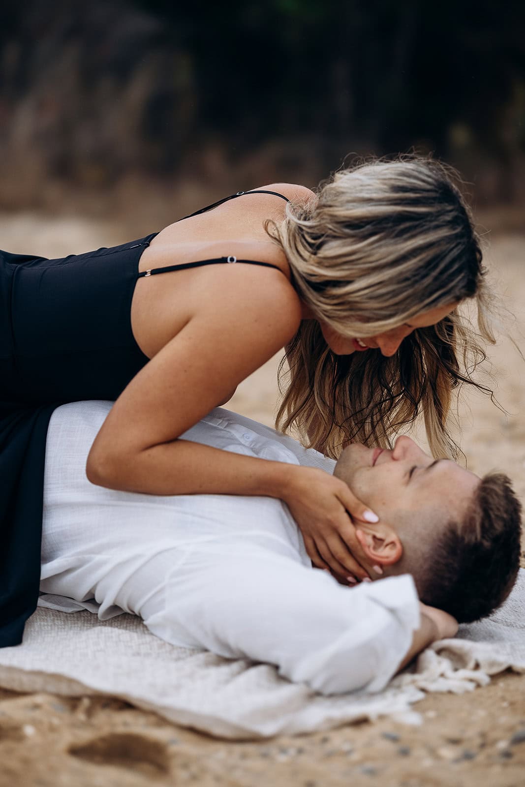 Woman in black dress leaning over man in white shirt lying on a blanket on sandy beach, sharing an intimate moment.