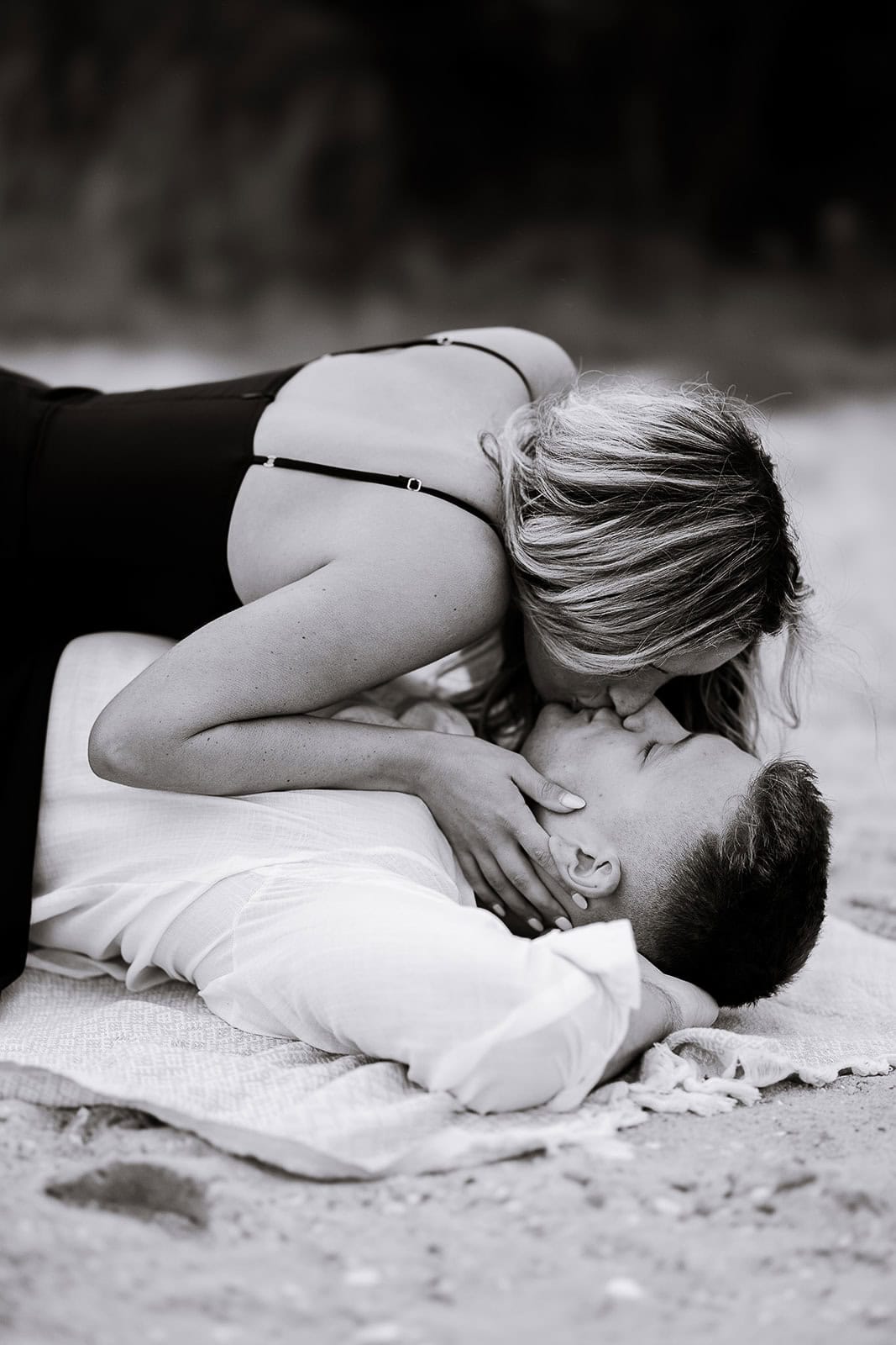 Black and white photo of a woman in a black dress leaning over a man in a white shirt lying on a textured blanket, sharing a kiss.