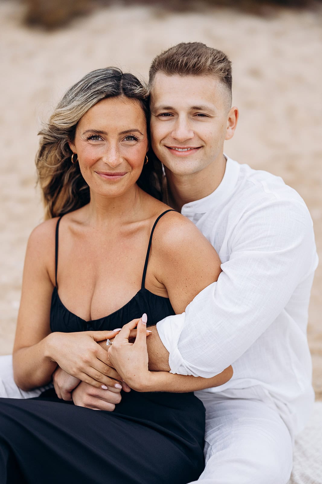 Close-up portrait of a couple sitting closely, the man in a white shirt embracing the woman in a black dress, both smiling gently against a soft, blurred background.